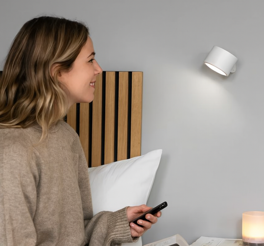 Woman sitting on a bed using a smartphone with a wall-mounted light fixture in the background.